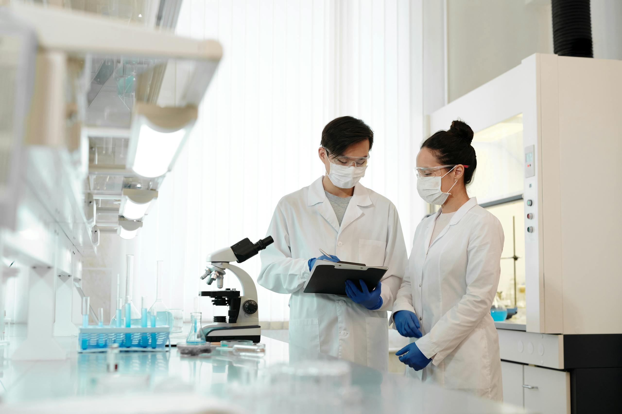 Two scientists in a laboratory wearing masks, discussing research with a microscope.